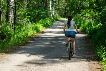 The girl rides a bicycle through the forest. Back view of the cyclist. A girl in shorts and a T-shirt rides a bicycle.