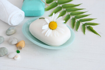 Fototapeta na wymiar White soap with a chamomile flower on top in a blue soap dish next to green leaves, with a towel and stones of the sea. Spa concept, treatments, body hygiene, natural cosmetics