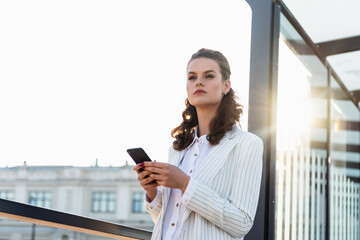 Fototapeta premium Smiling curly woman wearing trendy glasses walks down the central city street and uses her phone. Business woman using smartphone at commuter train station.