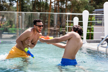 excited arabian man with friend fighting on water guns in pool