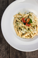 Four cheese penne pasta with basil and tomato in white plate on wooden table. Italian pasta. Pesto souce. Top view photo.