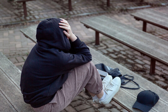 A Teenage Girl In A Black Hoodie With A Hood On Her Head Is Sitting On A Bench, Covering Her Face With Her Hands, Back View. Life And Problems Of Teenagers, Loneliness Concept