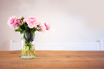 A bunch of pastel pink peony with green leaves in glass vase on the wooden table near white wall texture, cozy bright decoration for stylish house with copy space