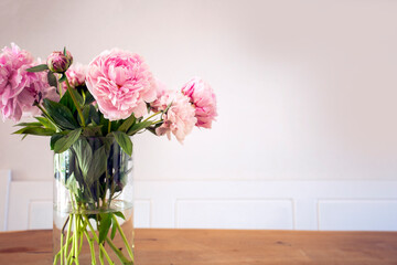A bunch of pastel pink peony with green leaves in glass vase on the wooden table near white wall texture, cozy bright decoration for stylish house with copy space