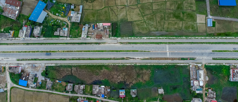 Aerial view of six-lane highway passing through meadows, fields and scattered houses towards mountains in countryside Nepal.