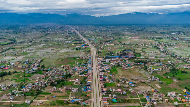 Aerial view of six-lane highway passing through meadows, fields and scattered houses towards mountains in countryside Nepal.