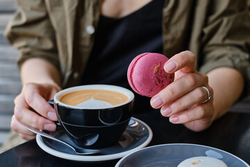 Woman with a cup of aromatic coffee in her hands sits on the summer terrace of the restaurant. The girl drinks hot latte after breakfast with sweet macaroon dessert. Delicious French dessert macaroon