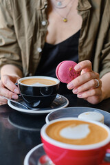 Woman with a cup of aromatic coffee in her hands sits on the summer terrace of the restaurant. The girl drinks hot latte after breakfast with sweet macaroon dessert. Delicious French dessert macaroon