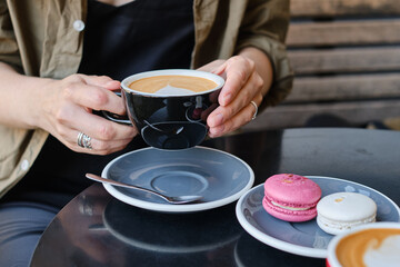 Woman with a cup of aromatic coffee in her hands sits on the summer terrace of the restaurant. The girl drinks hot latte after breakfast with sweet macaroon dessert. Delicious French dessert macaroon