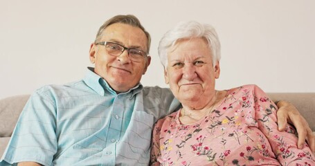 A long married couple, relaxing together at home on a sofa, smiling pensioners, a couple in love, a grandfather embracing a grandmother, looking at the camera, happy, cheerful