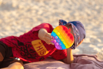 A little white boy on the beach sitting on the sand holds a finger toy Popit in front of him with hand. Trendy educational anti stress toy.