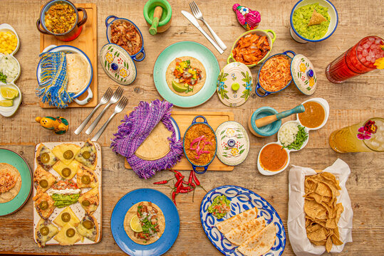 Top view image of traditional Mexican food on light wooden table. Forks, cochinita pibil in a pan, synchronized quesadillas, corn nachos, corn tortillas, chicken tinga, avocado guacamole