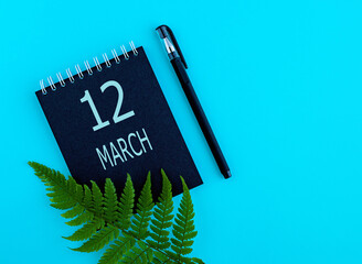 March 12th. Day 12 of month, Calendar date. Black notepad sheet, pen, fern twig, on a blue background. Spring month, day of the year concept