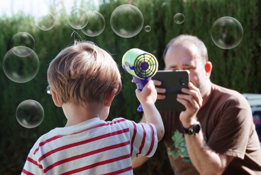 Man photographing his son playing with a soap bubble gun in the garden