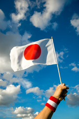Hand with Japan red and white wristband holding a Japanese flag waving in bright sunny blue sky - 442995331