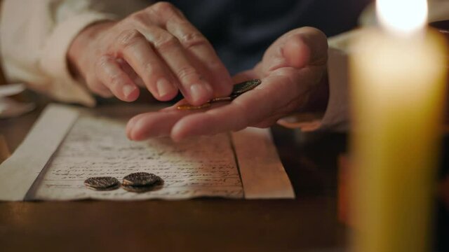 A close up candlelit scene of a Caucasian 18th century man counting and sorting various types of coins from his coin collection.