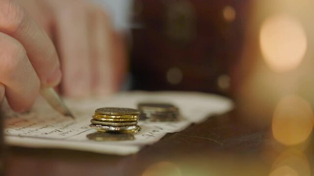 An close up 18th century scene lit by candlelight of a man documenting with a quill pen observations he has made about a stacks of coins.