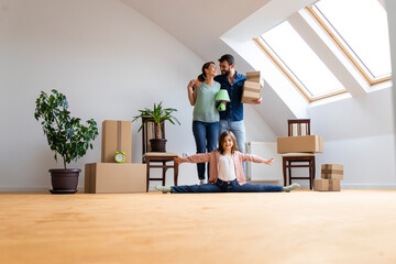 Fototapeta na wymiar Portrait of happy caucasian family moving in new apartment with flexible little girl doing gymnastics exercise and parents carrying belongings.