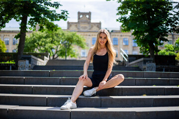 Young blonde sportive woman in a black tank top and in black tight sports shorts sits on the stairs