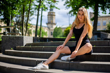 Fototapeta na wymiar Young blonde sportive woman in a black tank top and in black tight sports shorts sits on the stairs