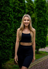 Young beautiful sporty blonde woman in a black T-shirt and in black tight sports shorts in good shape posing in front of green trees