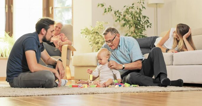 Family happy, satisfied looks at child playing on the carpet with wooden blocks, toddler is the center of attention, grandfather devotes time to grandson, kisses him on the head, takes care