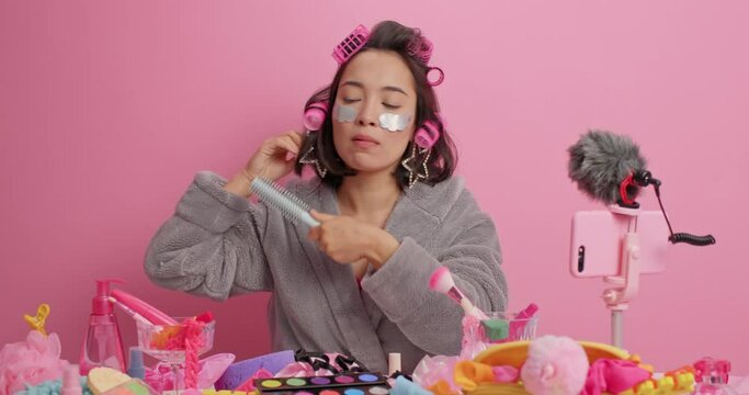 Funny Asian lady applies beauty patches under eyes sings song holds comb near mouth as if microphone makes hairstyle with rollers dressed in bathrobe records video blog sits at table with cosmetics