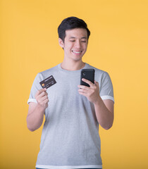 Excited Asian man holding credit card and smartphone feeling excited while looking at mobile phone isolated on colour background