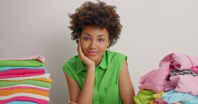 Serious curly haired Afro American woman in green blouse sits at table folds laundry at home looks with calm satisfied expression at camera isolated over white background. Household concept.