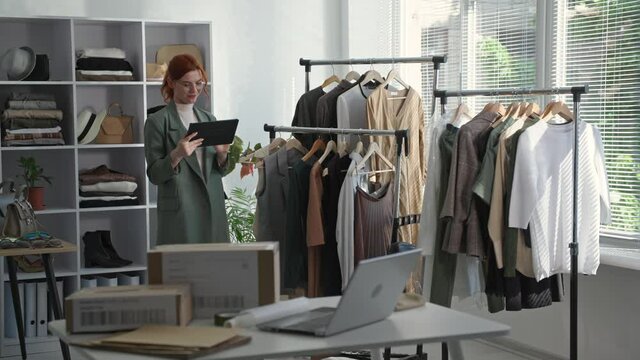 retail store, a young female entrepreneur holding a tablet in hand takes an inventory of clothes on the shelves in a boutique