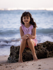 Fototapeta na wymiar Young girl sitting on the rock near the ocean. Smiling girl. Happy childhood. Spending time on the beach. Vacation in Asia. Pandawa beach, Bali, Indonesia