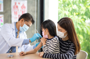 Asian  senior doctor wearing gloves and isolation mask is making a COVID-19 vaccination in the shoulder of child patient with her mother at hospital.