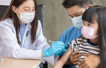 Asian  senior doctor wearing gloves and isolation mask is making a COVID-19 vaccination in the shoulder of child patient with her mother at hospital.