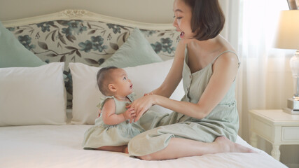 asian woman mother parent encourage baby infant to sit and crawl on soft bed at luxury home. 6 months kid put effort  learning lesson development skill. mom playing with baby infant on bed in bedroom.