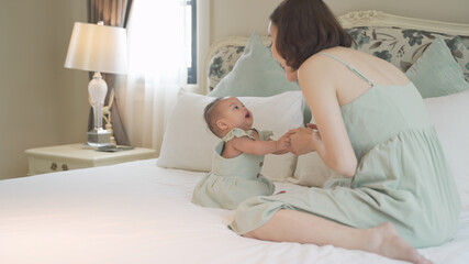 Obraz na płótnie Canvas asian woman mother parent encourage baby infant to sit and crawl on soft bed at luxury home. 6 months kid put effort learning lesson development skill. mom playing with baby infant on bed in bedroom.