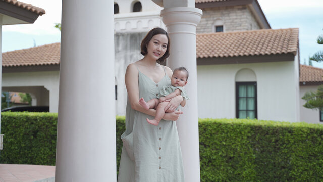 portrait of asian woman mother holding baby infant on balcony terrace at luxury home looking away the morning view. happy smile 30's female parent and 6 months baby at home near garden daytime.