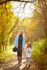 Naklejka na ściany i meble Mother and young little girl with blonde hair in an autumn park on a yellow and orange leaf background. Family walking in forest