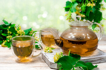 Glass teapot and two cups of tea with a linden tree on a wooden table on a blurred background and linden leaves, flowers. Healing tea.