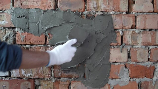 Close-up of the hands of a master builder in gloves, he holds a spatula and smears it with plaster on a brick wall.