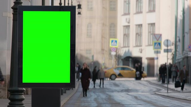 Modern empty billboard with a green screen for advertising on the busy street with defocused pedestrians at sunrise.