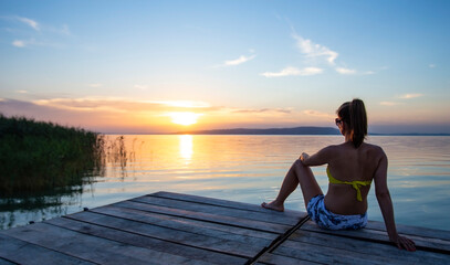 Lonely woman sitting on the seashore on a pier. Silhouette of young beautiful girl sitting on the beach next to reeds at sunset, viewing the sun. Blue orange yellow sky with some clouds Clam lifestyle