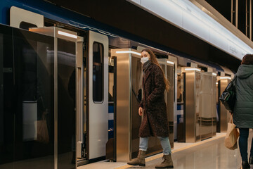A full-length photo of a woman in a medical face mask to avoid the spread of coronavirus who is entering the modern subway car. A girl in a surgical mask is keeping social distance on a metro station.