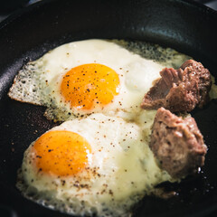 Men's breakfast - Two eggs are fried in a cast iron skillet with chunks of stew meat. Close-up