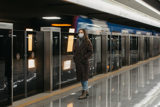 A full-length photo of a woman in a medical face mask to avoid the spread of coronavirus who is waiting for an arriving train on the subway platform. A girl in a mask is keeping social distance.