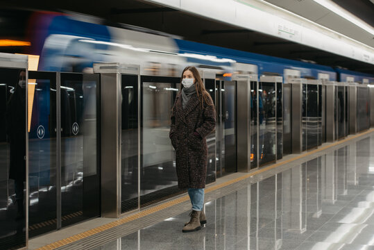 A full-length photo of a woman in a medical face mask to avoid the spread of coronavirus who is waiting for an arriving train on the subway platform. Girl in a surgical mask is keeping social distance