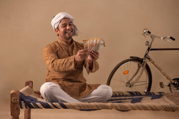 A TURBANED RURAL MAN HAPPILY COUNTING MONEY WHILE SITTING AT HOME