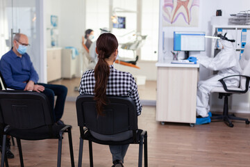Woman patient in dentistiry office waiting for diagnosis in dental clinic corridor. Dentist and staff dressed in ppe suit consulting patient. Filling health form.