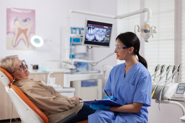 Dental assistant taking notes on clipboard in dentist clinic during elderly woman check up. Senior...