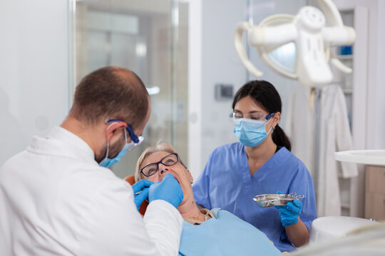 Stomatolog with assistant putting dental seal on senior woman teeth. Elderly patient during medical examination with dentist in dental office with orange equipment.