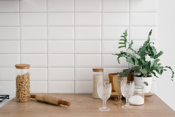 A glass jar with pasta, glasses, a rolling pin and a houseplant stand in the bright kitchen. Kitchen utensils, Scandinavian style